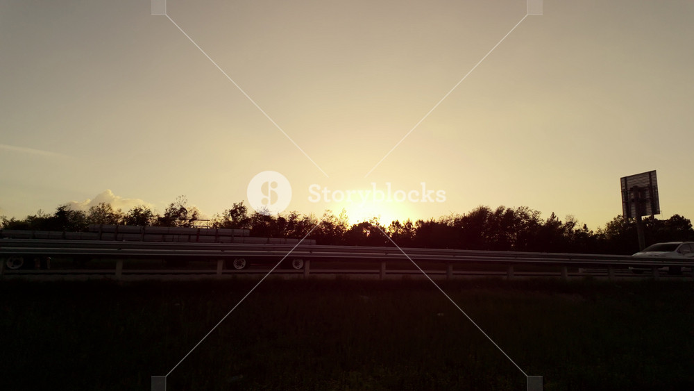 Trees and cars by fast as seen from the driving car. Soft gold sunset sunlight in the dark background. POV view of Sun set landscape