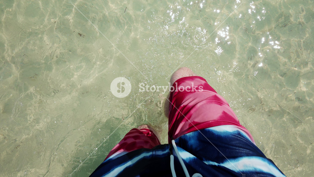 POV shot of a person standing barefoot in clear shallow seawater, with gentle waves moving around the legs, capturing a calm tropical beach