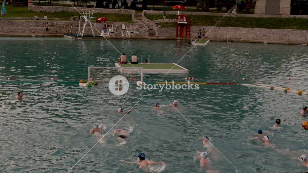 People playing water polo in an outdoor swimming area with floating goals and spectators. Summer sports and recreation at a public aquatic