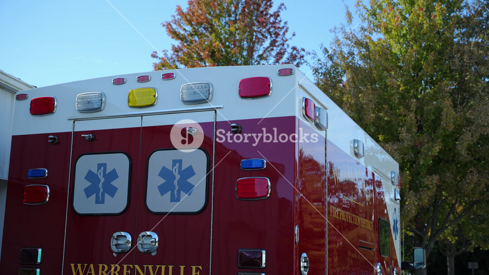 A red and white emergency ambulance, parked near a residential style garage or station building. USA IL Warrenville 2 OCT 2025