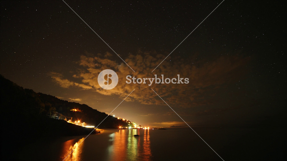 8K timelapse stars above harbor lights on calm sea, patchy clouds, small boats at dusk. Night sky glitter across coastal port glow, ocean reflections, drifting haze, moored vessels, shoreline view.