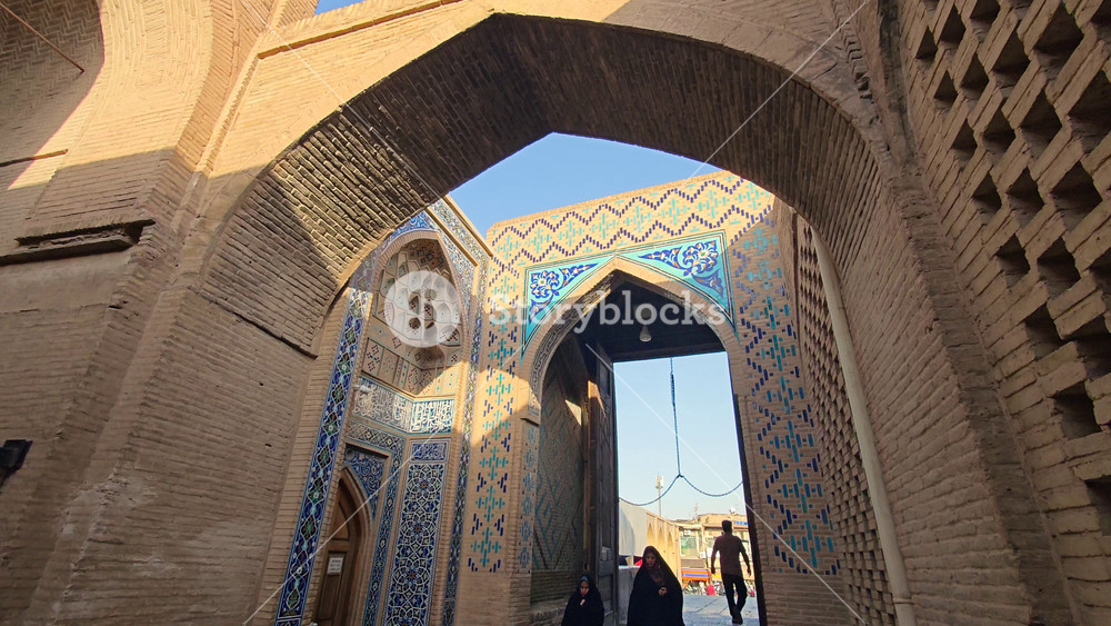 8K Iranian Muslim women wearing black chador near historic mosque gate beside brick bazaar street. Persian female figures pass tiled madrasa portal along old market lanes amid Islamic architecture.