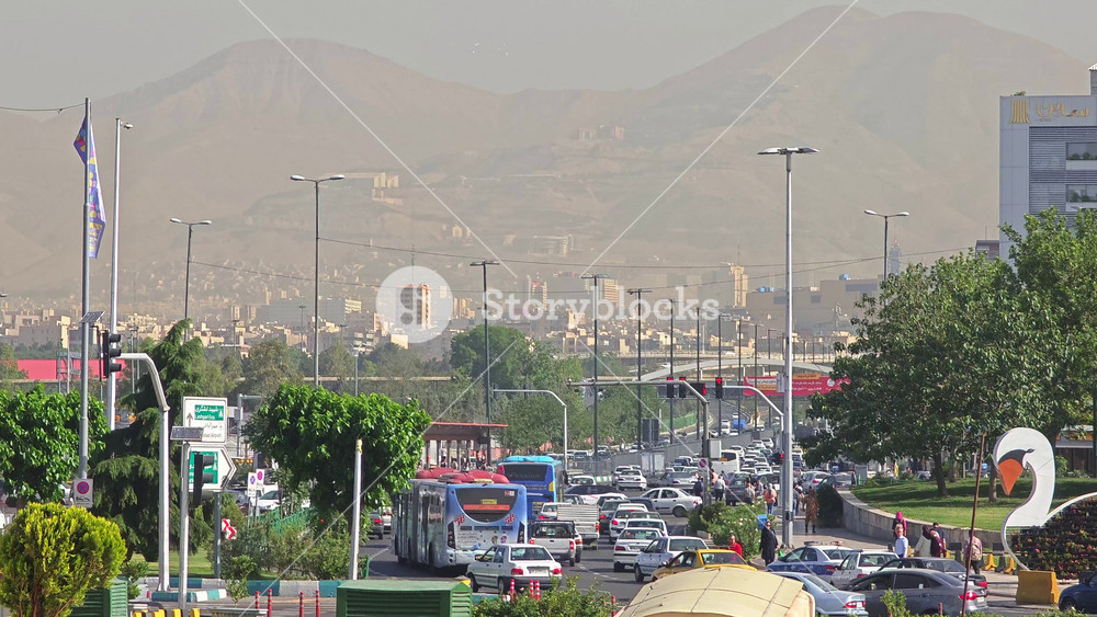 8K Tehran city park traffic with buses cars and Iranian flags in urban daily life scene. Metropolitan roadway beside green plaza showing commuters vehicles transit route plus civic banners.