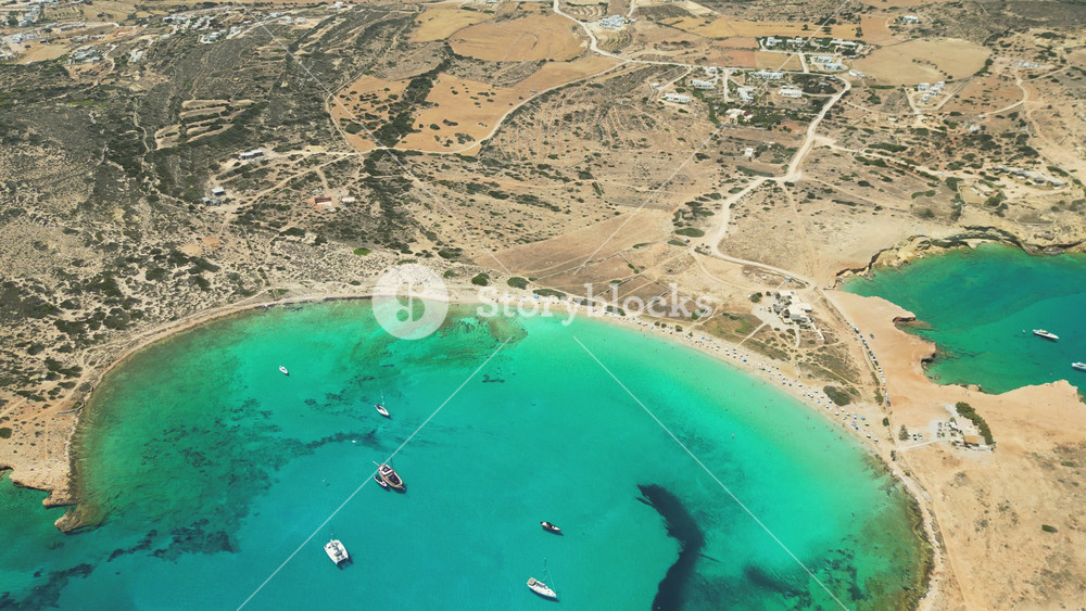 An aerial view of Koufonisia, Greece, showcases the clear turquoise waters of a peaceful bay. Sailboats add life to the serene landscape