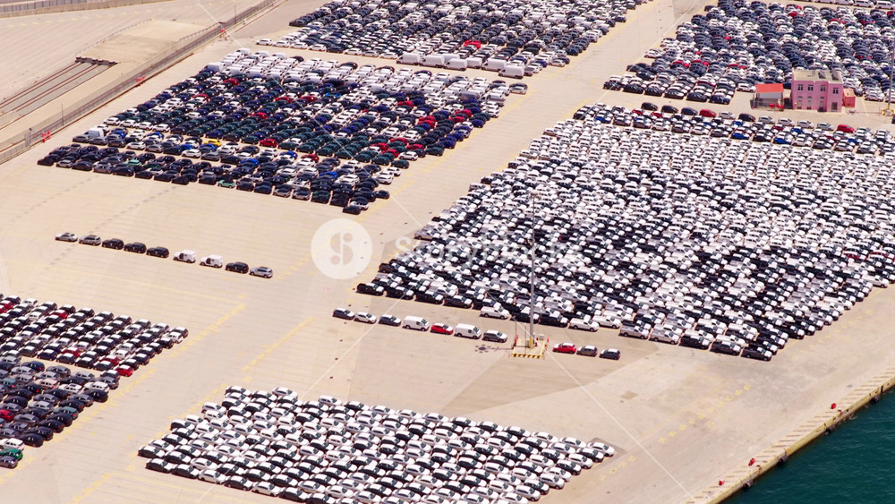 High-angle perspective showing thousands of brand-new cars perfectly parked in a massive lot at a shipping port, all prepared for export and distribution through the global supply chain