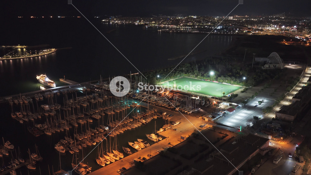 Stunning aerial footage showing a bustling city harbor filled with docked boats and a brightly illuminated soccer field, showcasing a vibrant coastal cityscape against the dark night sky