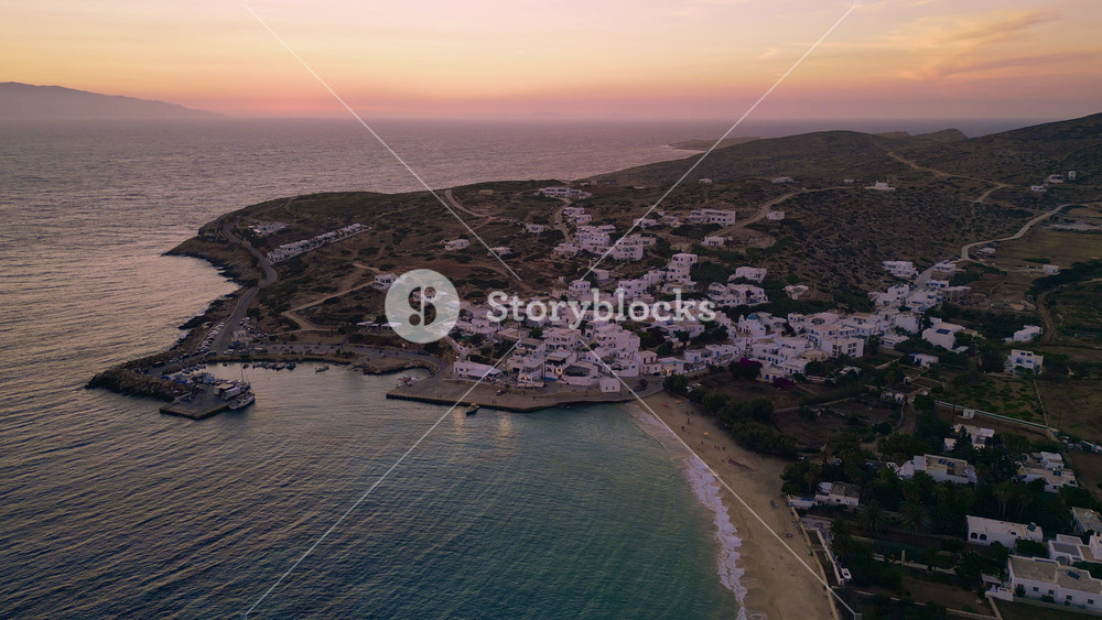 An aerial view of Stavros beach on the Greek island of Donousa as the sun sets. The whitewashed buildings of the village cling to the hillside above the beach.