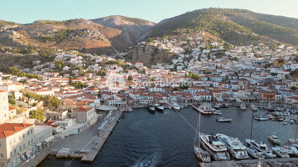 Breathtaking aerial drone footage of Hydra showcasing a steady low flight over small boats leaving the harbor at sunset. A picturesque view of the island's serene beauty.