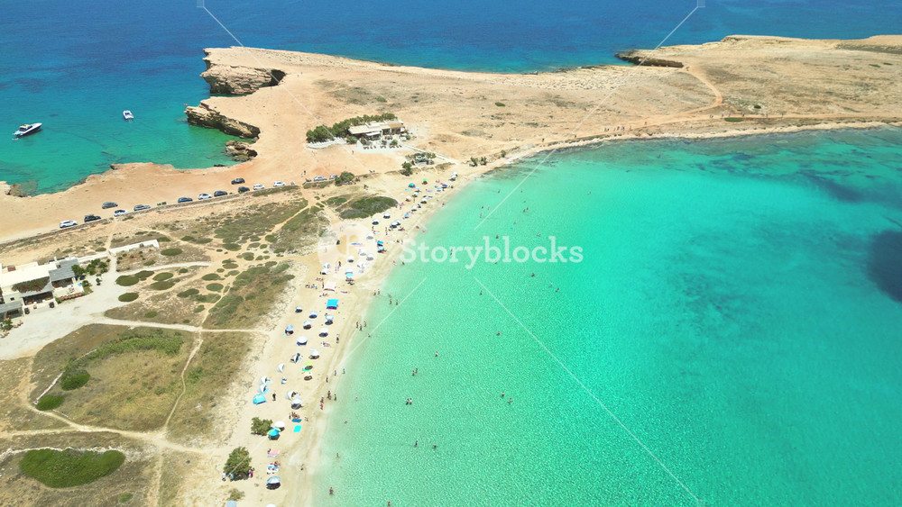 A breathtaking aerial view of Pori Beach in Koufonisia, Cyclades Islands, Greece, reveals crystal clear turquoise waters and a sandy shore dotted with sunbathers and umbrellas.
