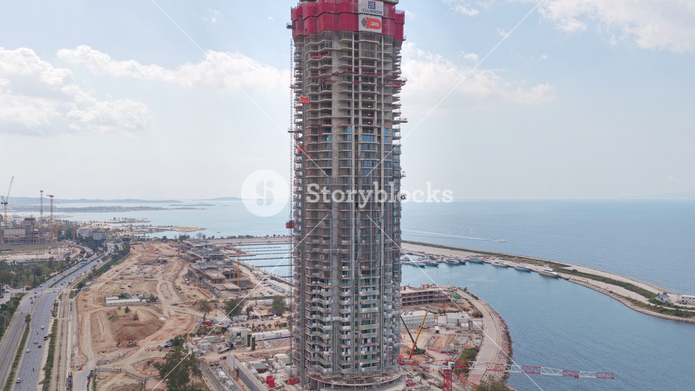 Aerial view of the riviera tower under construction at the ellinikon, with a boat sailing on the turquoise sea against the athens, greece cityscape on a cloudy day. Ellinikon, Athens Greece, July 21 2026.