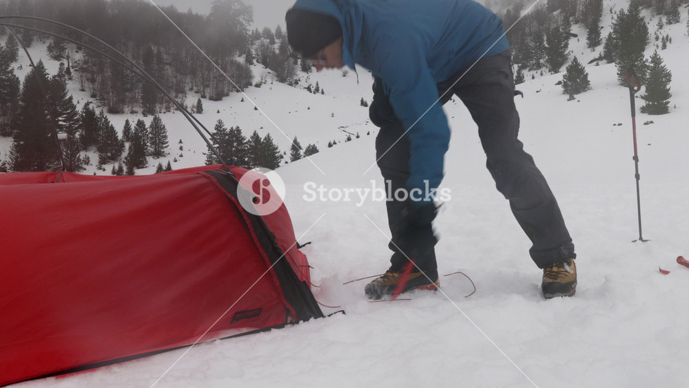 A person sets up a bright red tent in a snowy landscape. Snow falls heavily, covering the ground and creating a winter wonderland. Visibility is reduced in the frosty conditions.