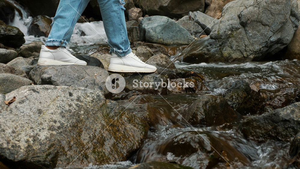 Close up of girl's legs in jeans and sneakers against mountain river in the Alps. Summer vacation and tourism concept.