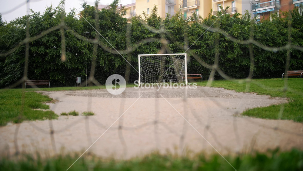 Detailed view through soccer net on rainy day showcasing deserted urban football pitch with lush green grass. Refreshing and calming outdoor sports scene.