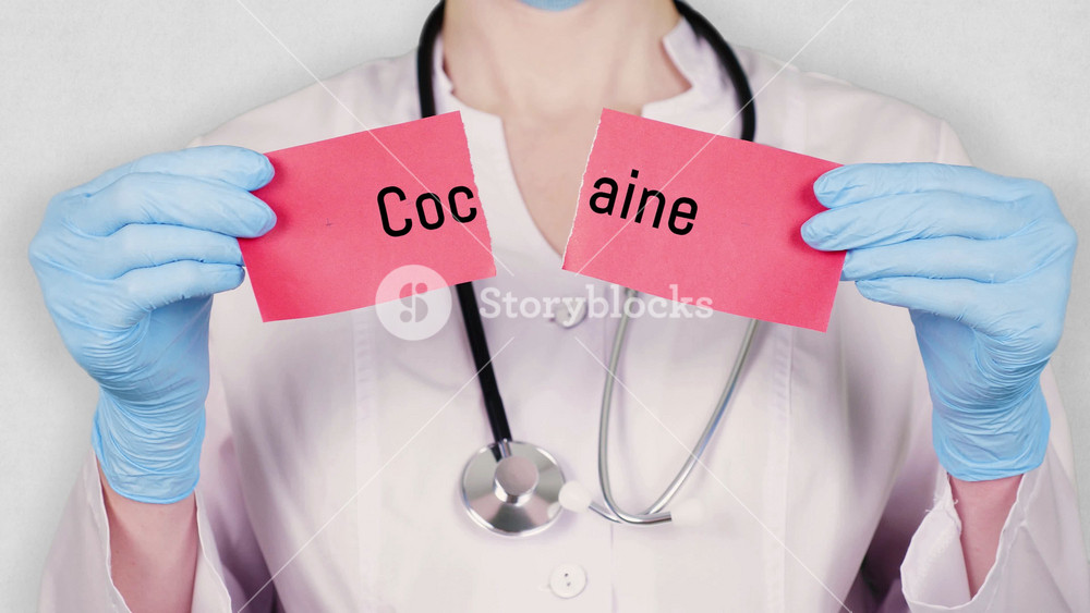 closeup, hands in blue medical gloves hold and tear red paper card with an inscription cocaine. doctor dressed in white medical uniform, has a stethoscope.