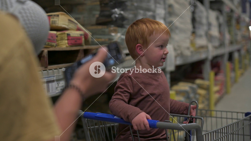 Young boy with red hair sits in shopping cart while adult holds smartphone, surrounded by shelves of building materials in a well-lit store