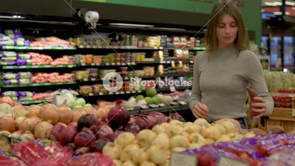 Woman on grocery store selecting onion from colorful assortment of red, white, and yellow onions displayed on grocery store produce bin with packaged potatoes vegetables in background grocery store