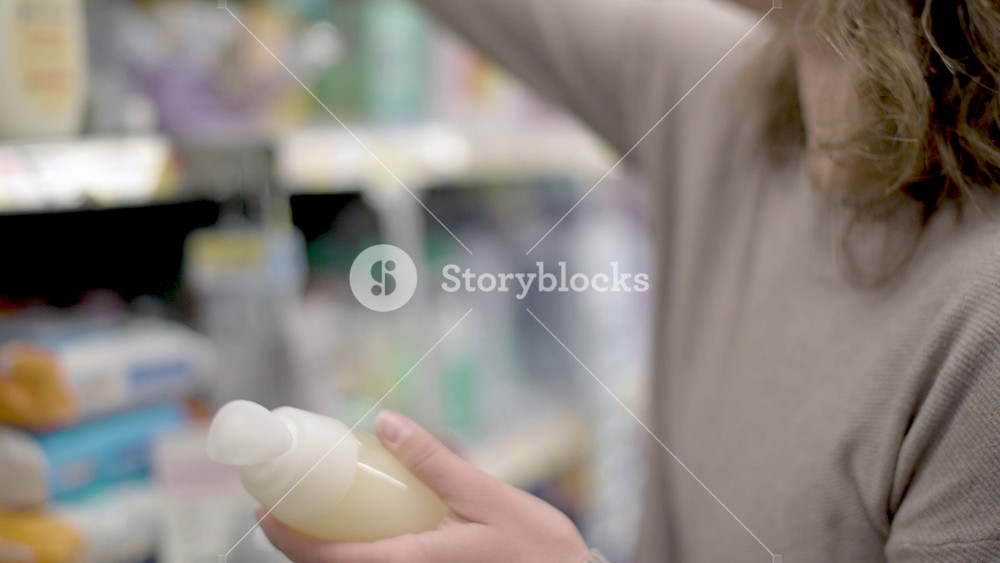 Woman in long sleeve shirt holding bottle of personal care product while reaching for another item from shelf in a brightly lit store aisle, browsing health and beauty supplies during shopping trip