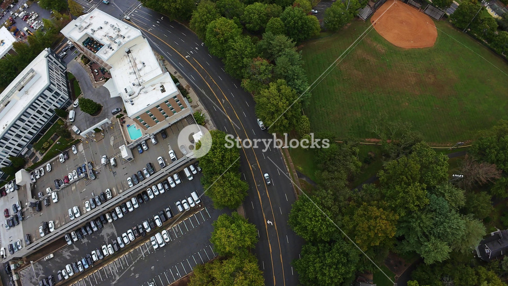 Aerial Asheville Ballfield Beside Commercial Parking Lot, Curved Road Frames Green Diamond Infield, Adjacent Buildings With Rooftop Pool