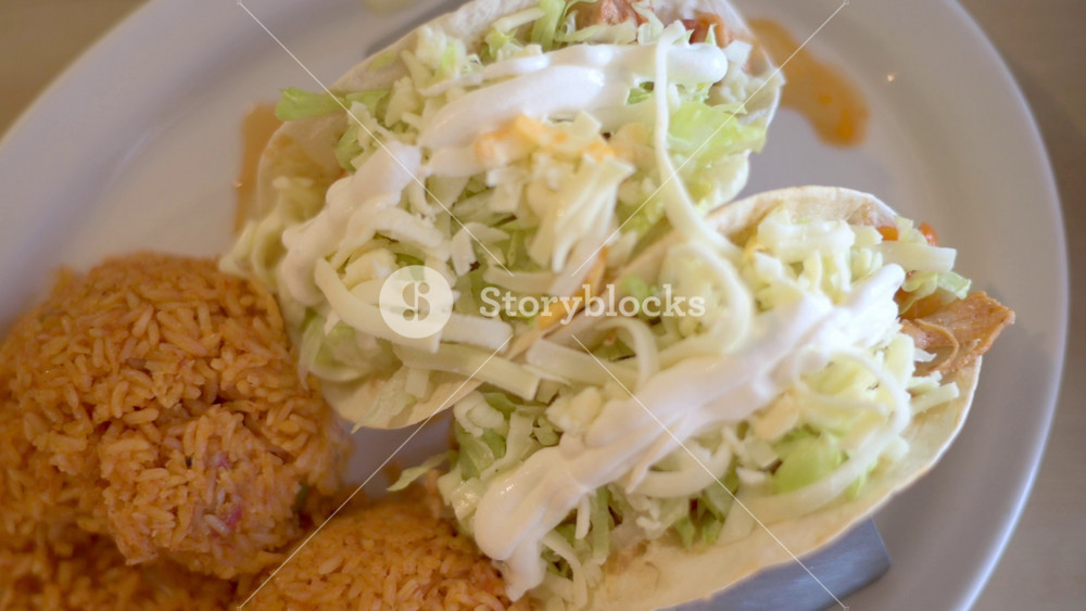 Close-up of plate with two soft tacos topped with shredded lettuce and white sauce, served alongside two scoops of seasoned Mexican rice on white plate, representing fresh and flavorful meal.