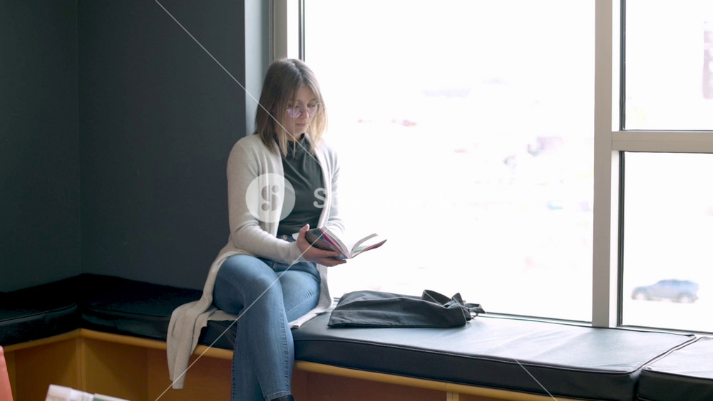 Asian woman with shoulder-length hair in white cardigan and jeans sitting on window bench flipping through book pages, natural daylight streaming in, casual and relaxed atmosphere