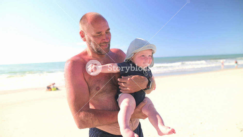 Caucasian man holds smiling baby in blue sun hat at beach, with gentle waves and sandy shore in background, showcasing joyful moments of summer life