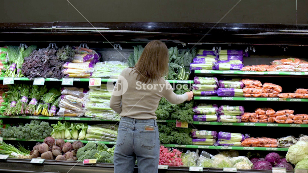 Woman standing in front of grocery store produce section reaching for leafy greens with shelves full of colorful vegetables including carrots, lettuce, beets, radishes, and cabbage grocery store