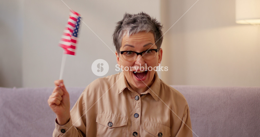 Latin woman waves small American flag cheering for national sports team at home. Senior lady makes victory gesture celebrating success of