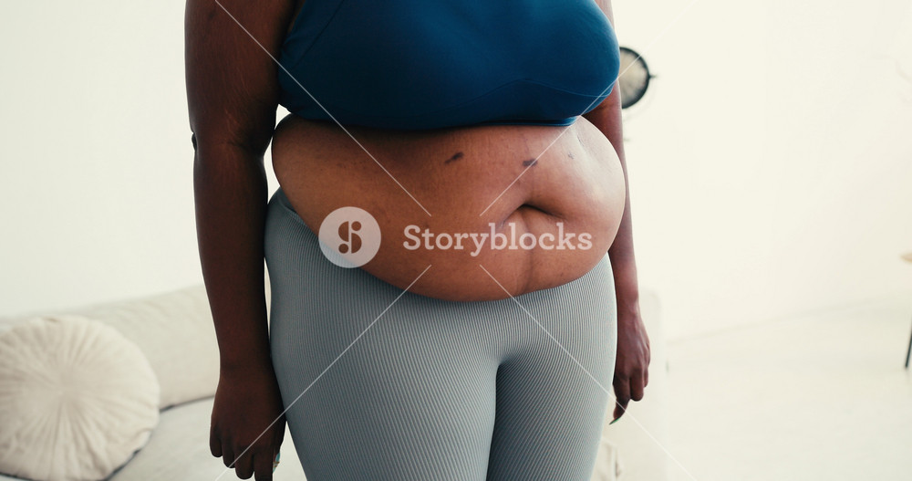 Overweight black woman reveals belly fat showing heart with hands. Female highlights body positivity and self acceptance standing in living