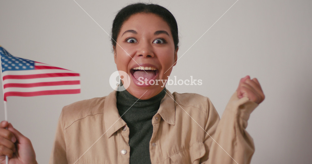 Ecstatic Hispanic woman waves small national flag of America on light background. Joyful brunette female celebrates USA Independence day