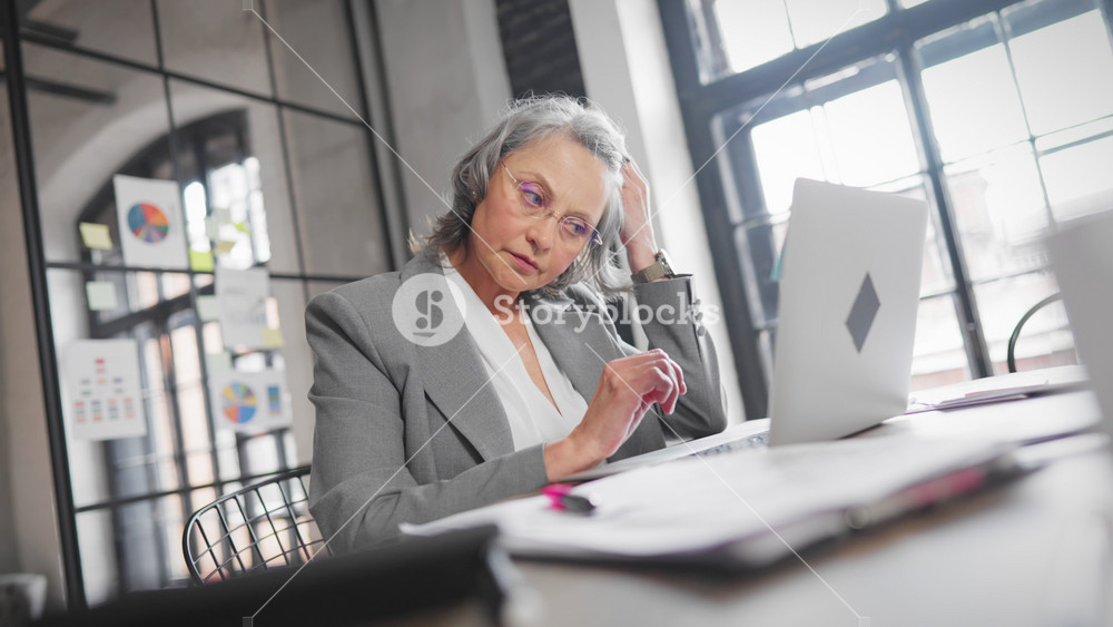 Medium portrait of tired mature woman employee in office, lady taking off. Senior female person using laptop, looking at display and bridge