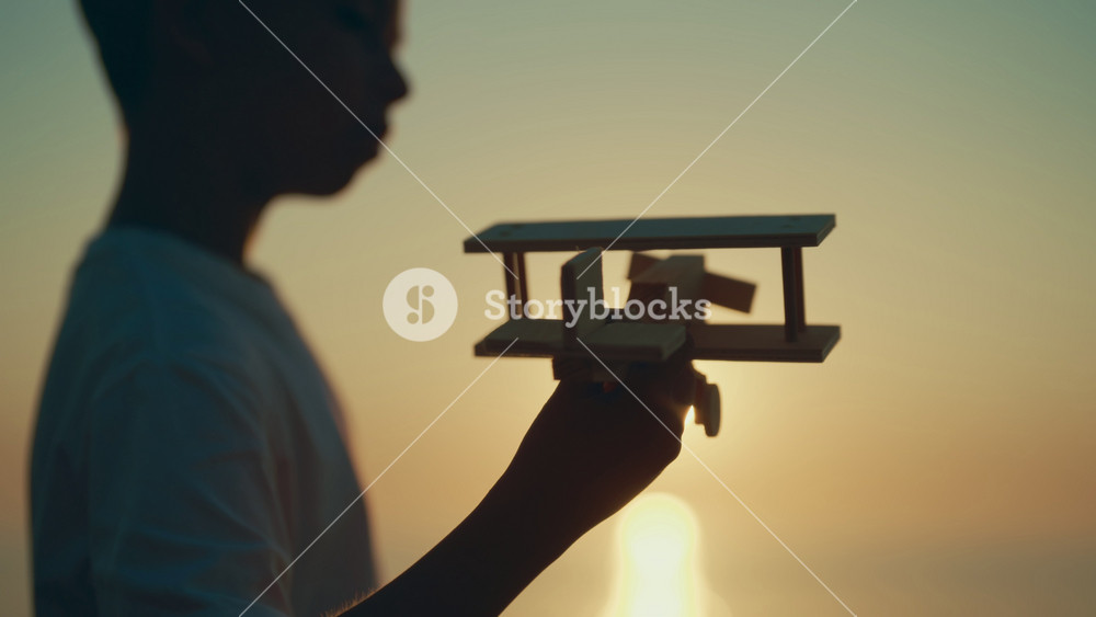 Silhouette of little boy with handmade model of aircraft in hands against sea. Amazing sunset above ocean, happy child dreaming about