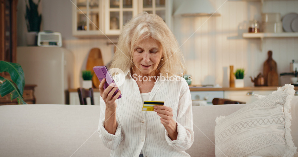 Retired woman talking her credit card data to phone scammers, internet fraud. Portrait of 70-years-old lady in cozy home interior, victim of