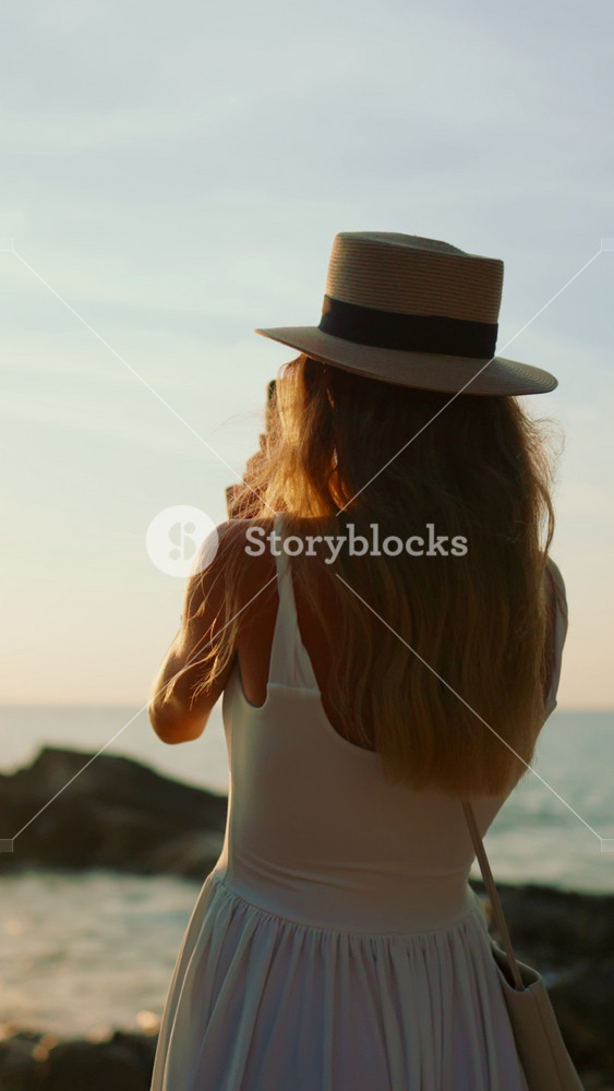 Pretty woman admiring seascape from shore in sunset or sunrise, travel to sea in summer vacation. Vertical back view of carefree slender