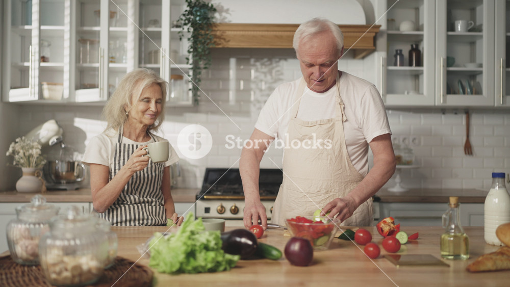 Nature man cooking salad for his wife, old lady admiring and relaxing with tea. Portrait of elderly spouses in home kitchen, happiness and