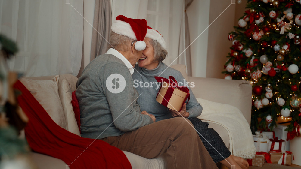 Aged man giving Christmas gift to his pretty wife in winter holidays, surprise. Celebrating New year and Xmas together at home, calm weekend