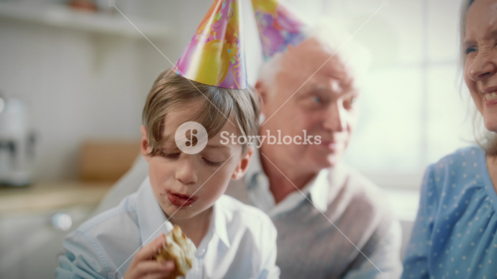 Caucasian little boy eating delicious birthday cake in merry family party. Portrait of preschooler with cap on head biting sweet tasty