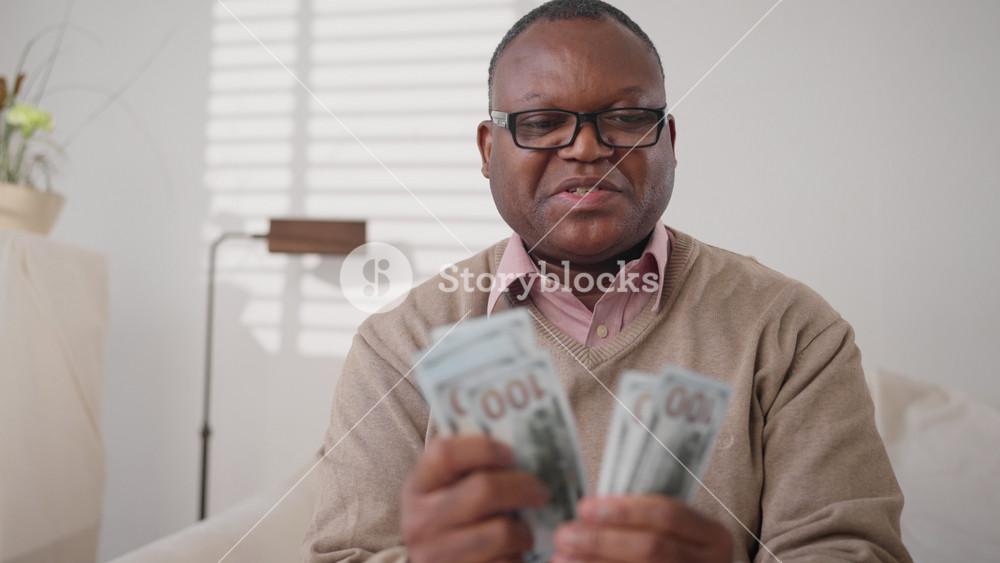 Happy middle-aged african american man counting new hundred dollars bills. Medium portrait of black male person in home interior, salary and