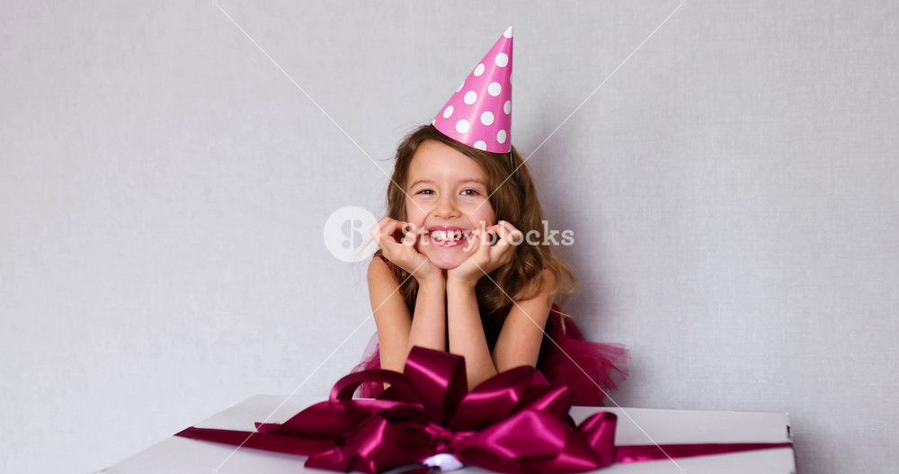 Portrait of joyful smiling little girl in pink dress and hat put her elbows on big present gift box, at home birthday party streamers, Happy birthday. Celebrating