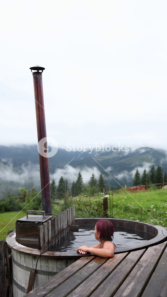 A teen girl relaxes in a hot vat while surrounded by mountains and trees. She enjoys her vacation in nature with cool weather and scenic views. The young girl looks content and happy.