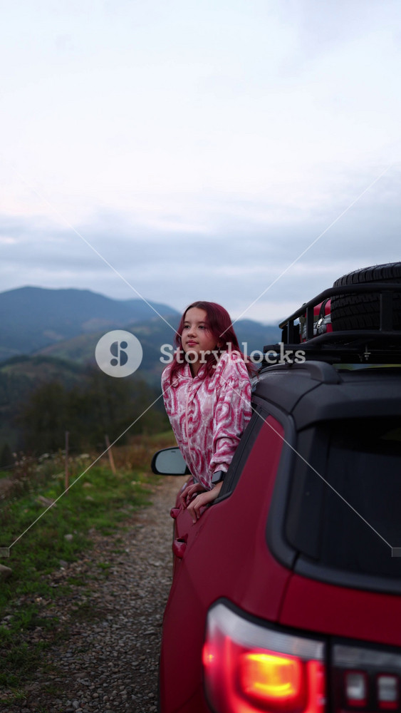A girl stands beside a car, taking in the view of mountains. The sky is filled with clouds, and the landscape shows rolling hills. This happens during late afternoon.