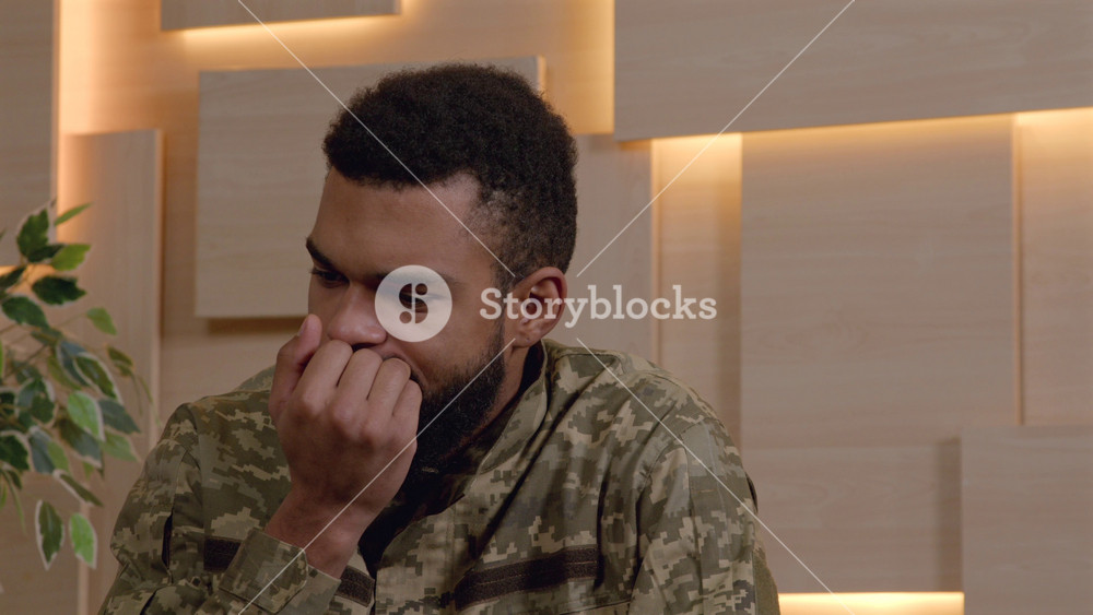 Portrait of worried uncertain black male army soldier in military uniform nail biting, expressing anxiety and tension while visiting counsellor during psychotherapy session office.