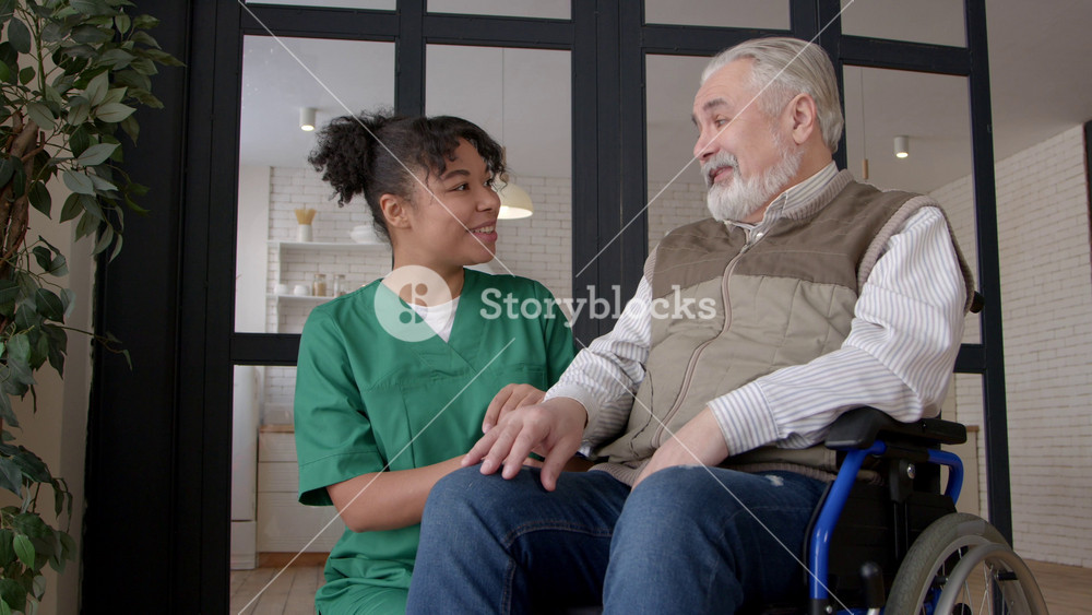 Cheerful senior handsome grey haired male wheelchair user and friendly pretty African American female caregiver in uniform enjoying leisure , communicating and sharing in care home.