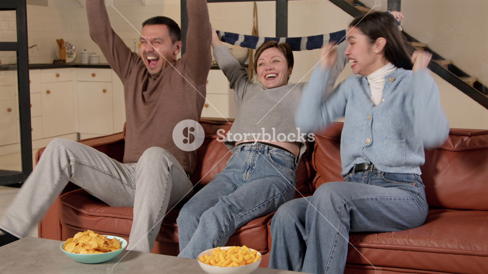 Joyful middle aged hearing impaired parents with American football ball and excited teenage daughter with hearing loss watching football game on tv, celebrating scoring drive and raising hands up.