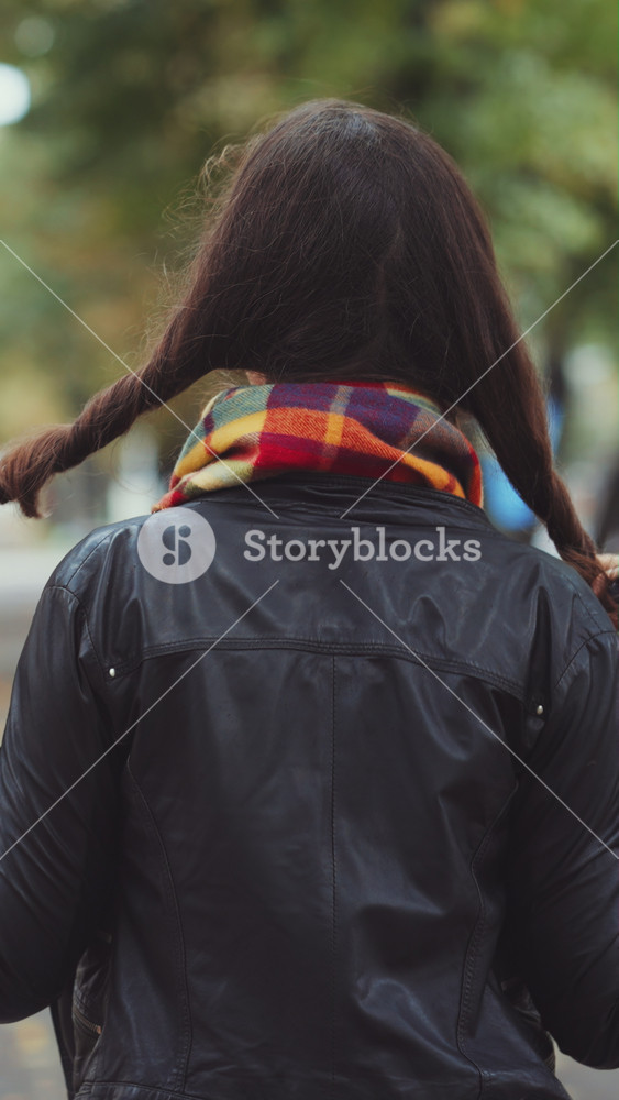 Back view of young woman in black leather jacket and checkered scarf. Brunette female walking in autumn park and touching hair on sunny day