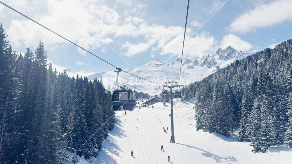 Scenic ski lift ride above snowy alpine slopes with skiers descending the mountain in Courchevel, France, featuring panoramic winter landscape and sunny blue sky in the French Alps.