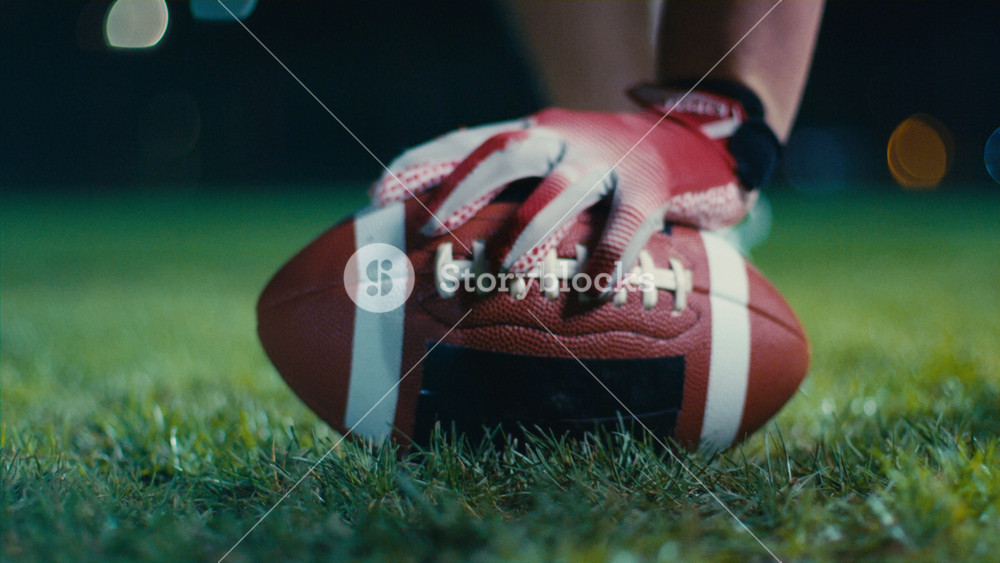 Close-up of an American football player wearing gloves, holding a ball on a field under bright stadium lights at night
