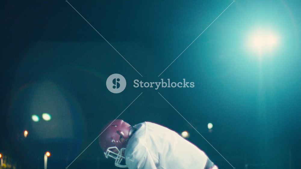 Portrait of an American football player at night at stadium lights with a helmet and protective gear uniform standing on one knee on the