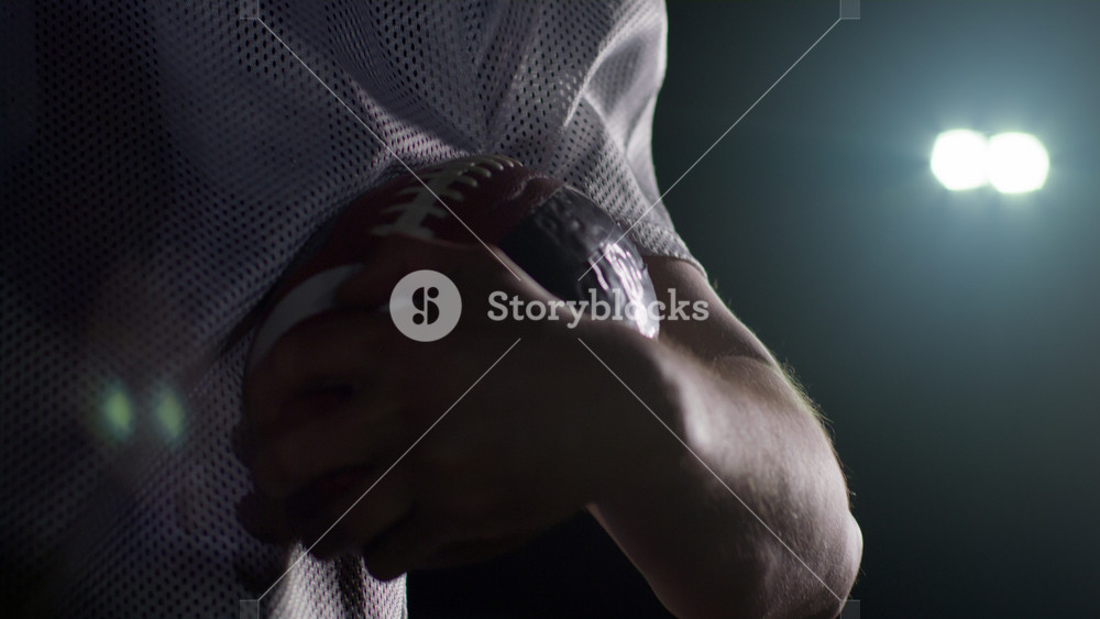 Portrait of a Caucasian male American football player wearing a team uniform, pads and a helmet, holding a football in his hands, raising