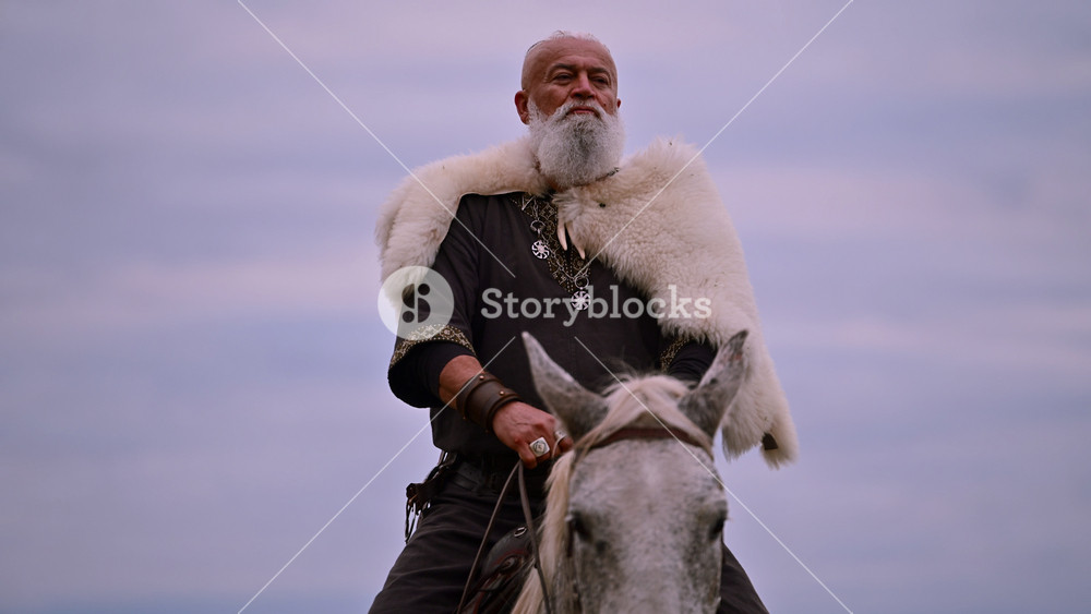 Elderly Viking warrior in fur cloak riding gray horse across open field at sunrise, mountains and distant cross behind him, moody cinematic