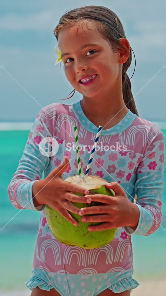 A close-up portrait of little girl drinking a fresh coconut cocktail with turquoise water on the shore of the Indian Ocean. A smile on the